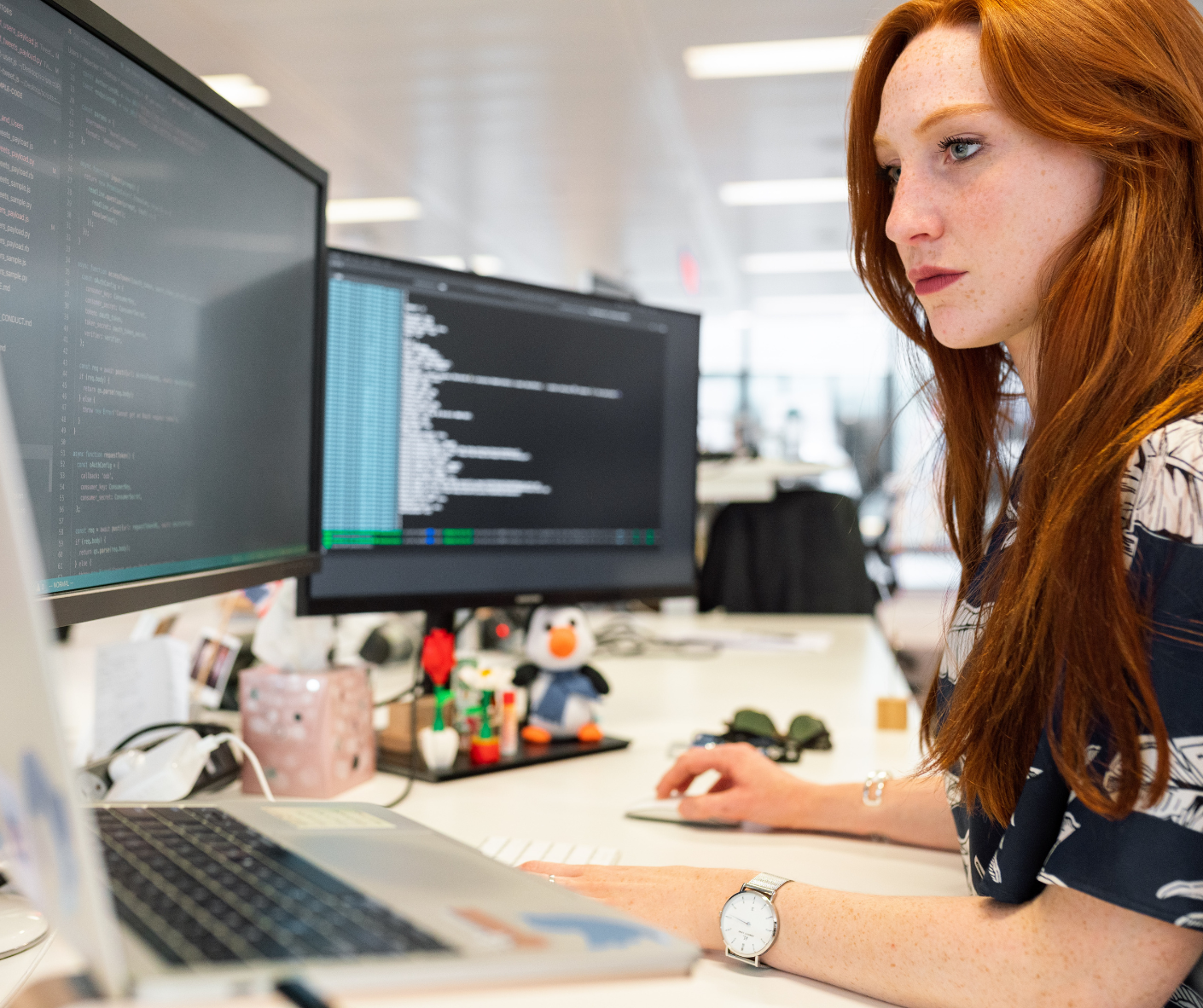 red-haired-woman-looking-at-3-monitors-on-work-desk